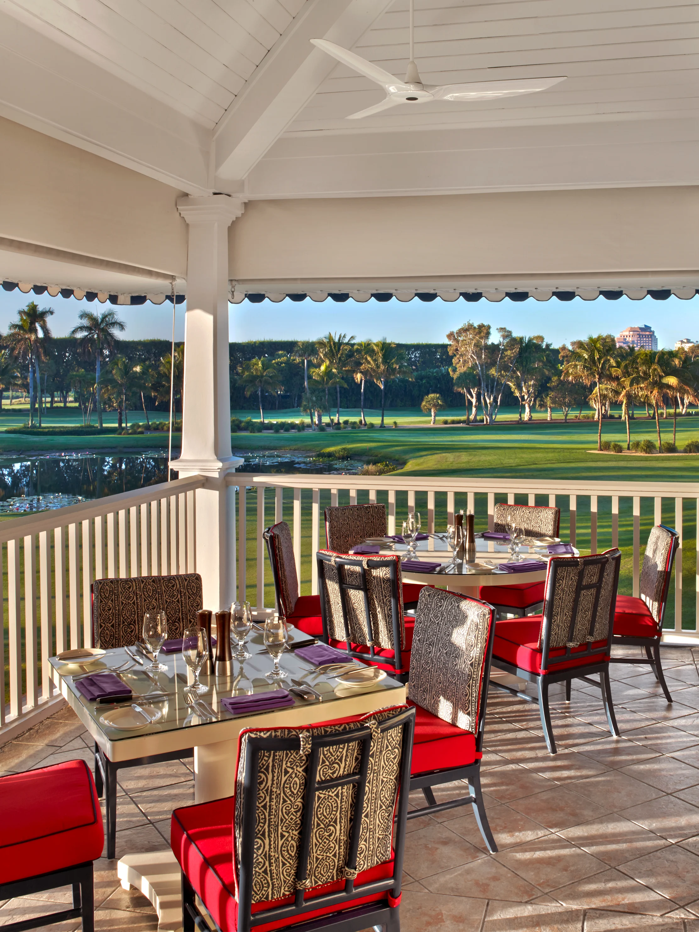 Covered patio dining area with red-cushioned chairs overlooking a lush golf course and palm trees.