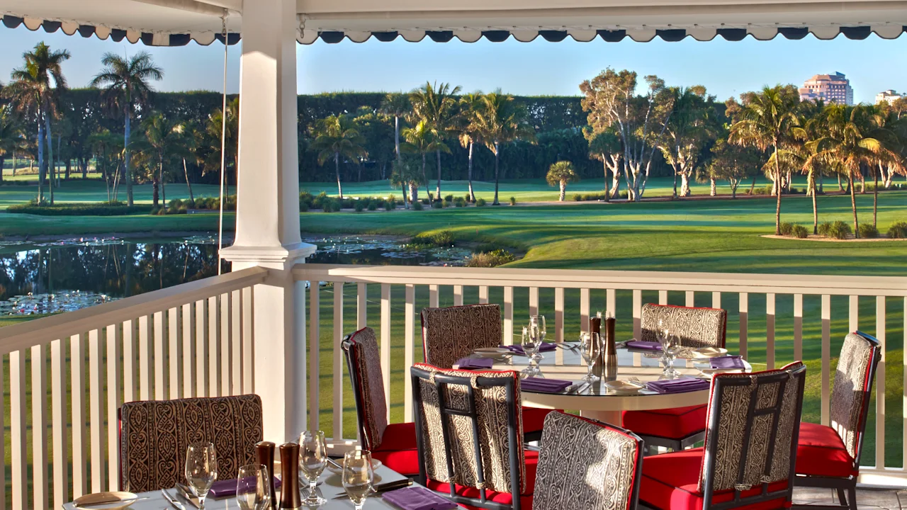 Covered patio dining area with red-cushioned chairs overlooking a lush golf course and palm trees.