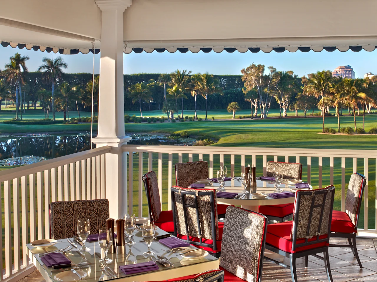 Covered patio dining area with red-cushioned chairs overlooking a lush golf course and palm trees.