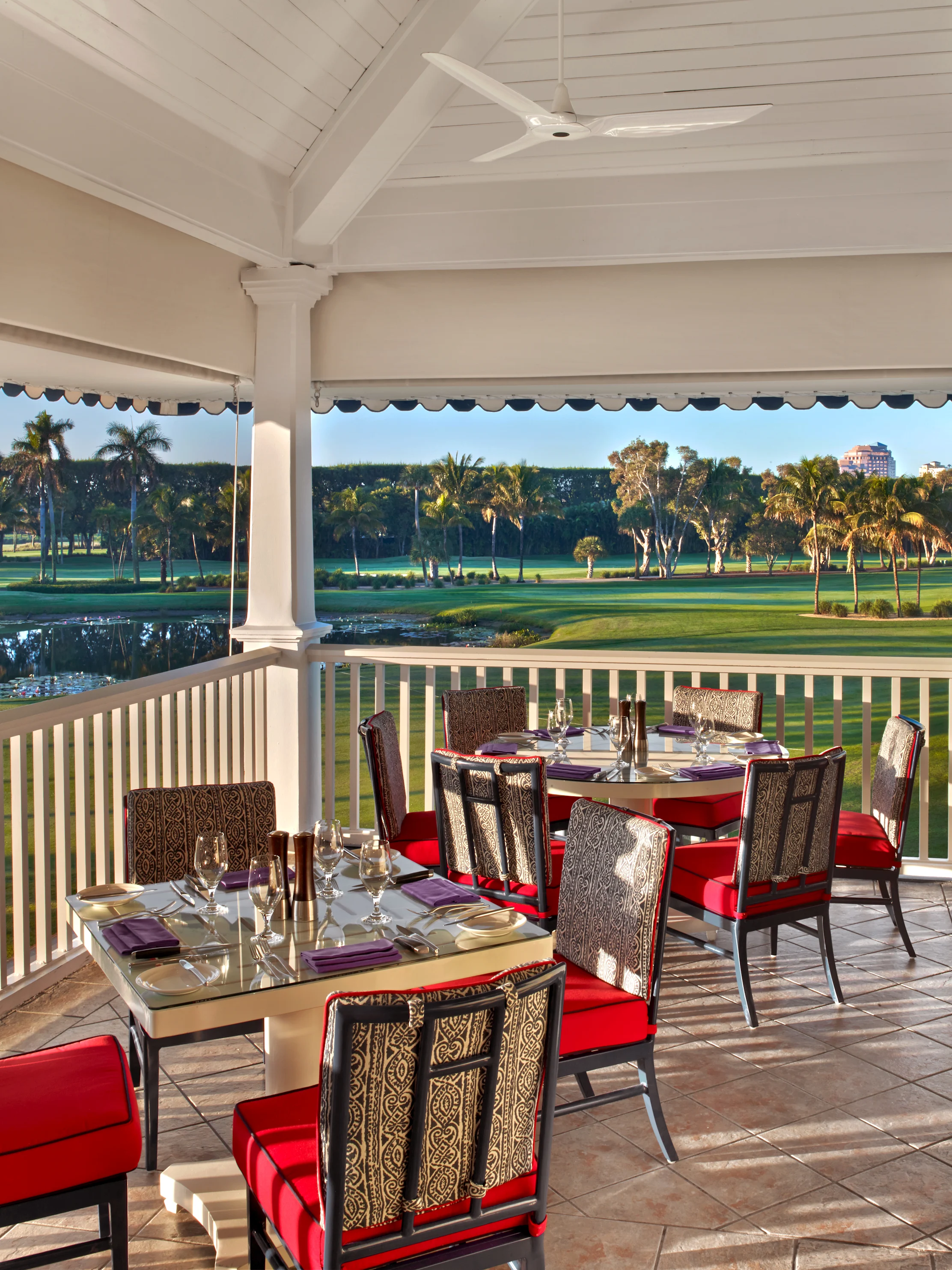 Covered patio dining area with red-cushioned chairs overlooking a lush golf course and palm trees.