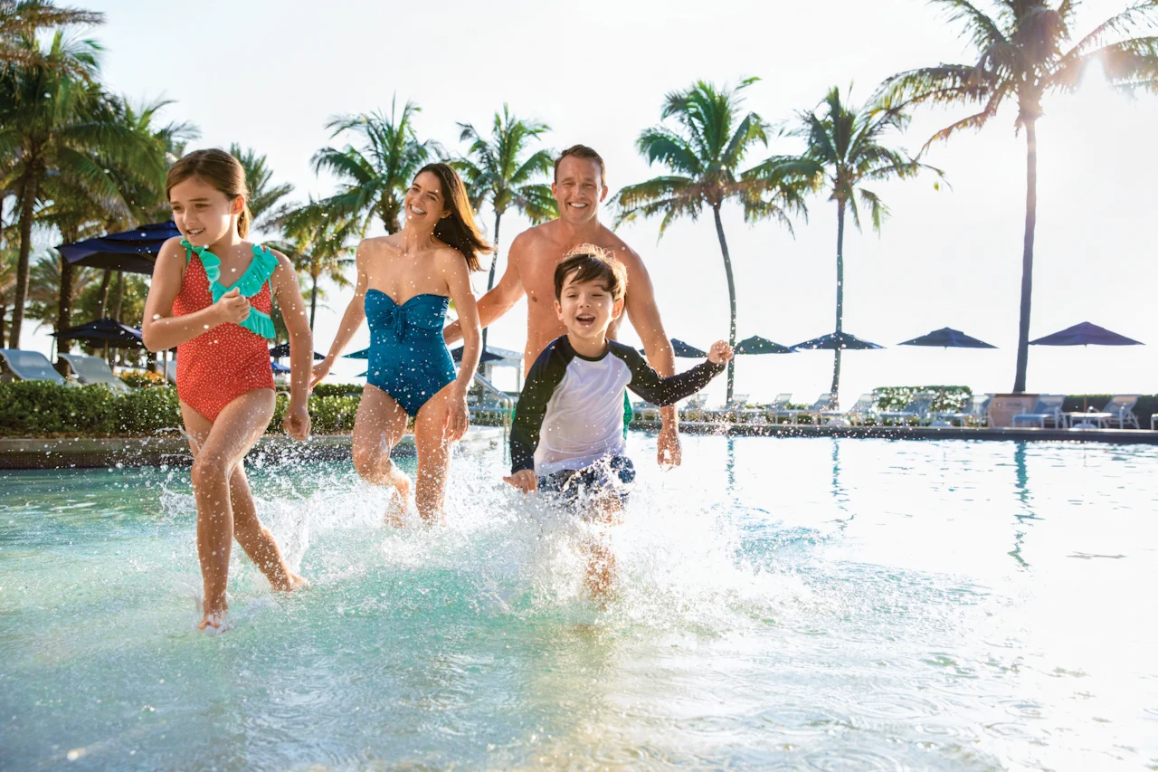 Happy family of four running through shallow resort pool with palm trees in background.