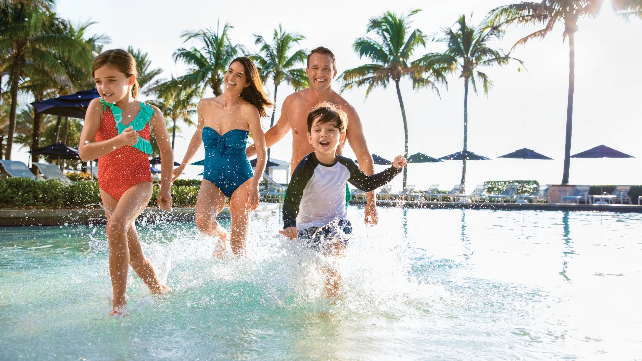 Happy family of four running through shallow resort pool with palm trees in background.