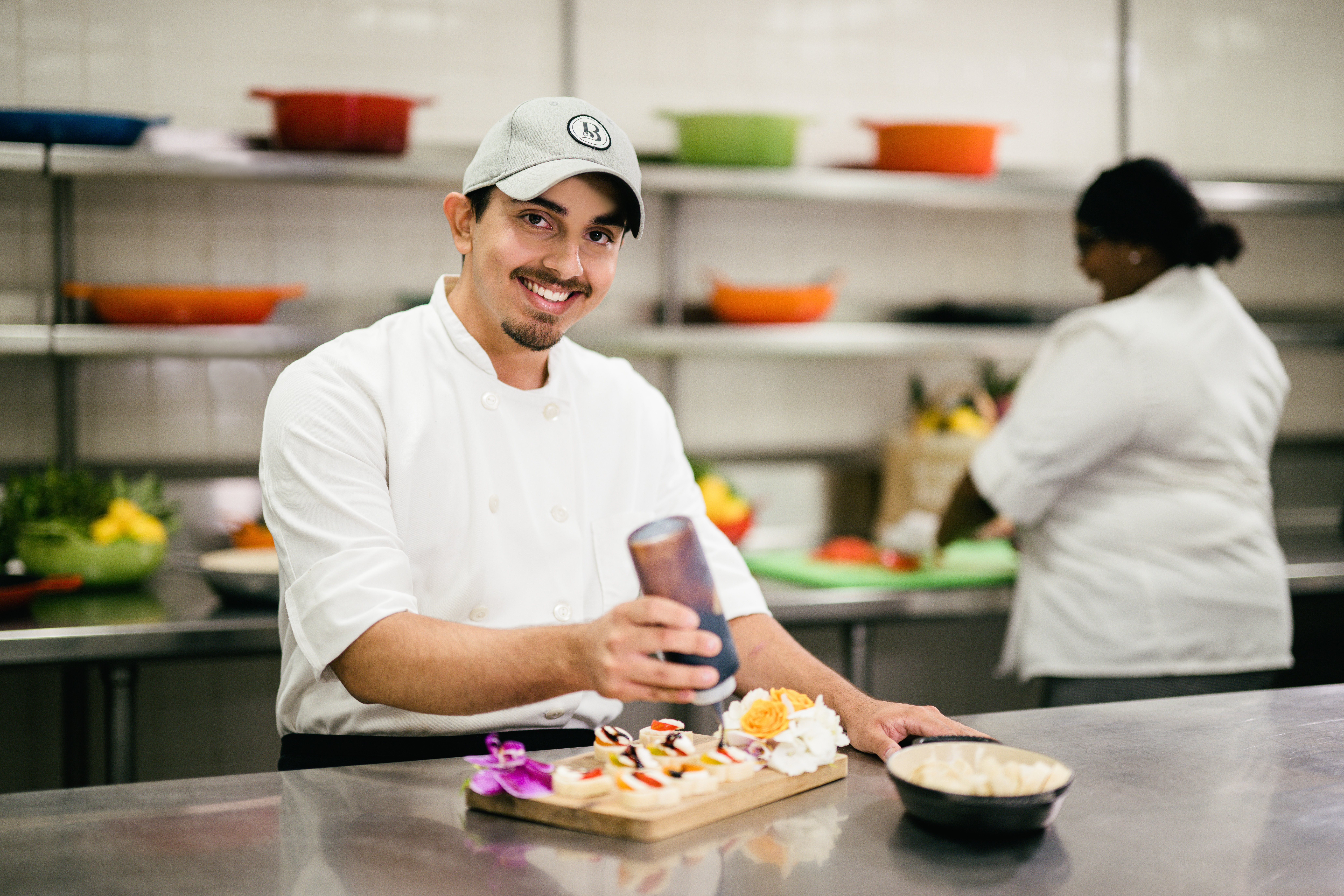 Smiling chef in white uniform and cap holding a drink in a commercial kitchen with colorful dishes.