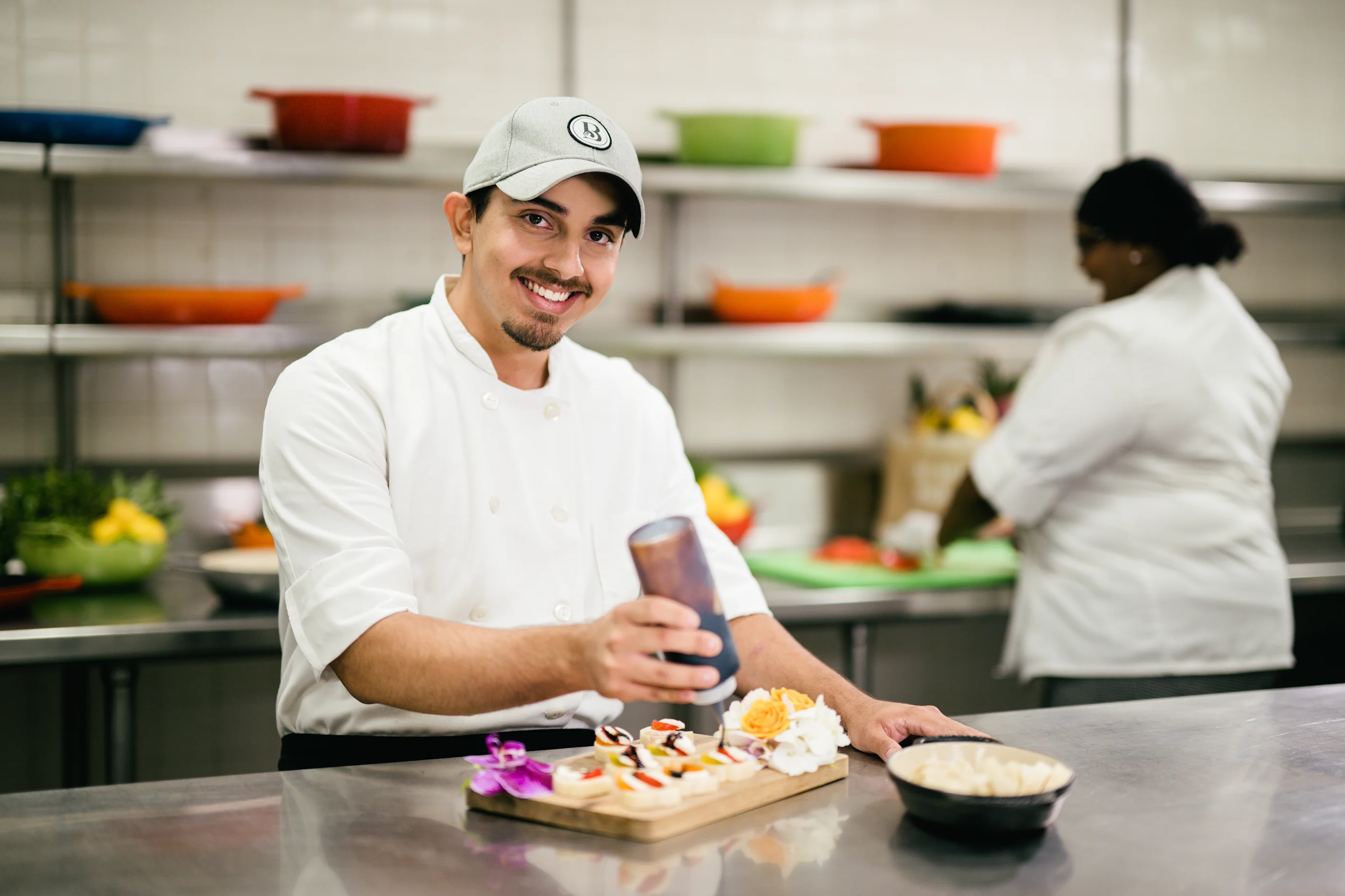 Smiling chef in white uniform and cap holding a drink in a commercial kitchen with colorful dishes.