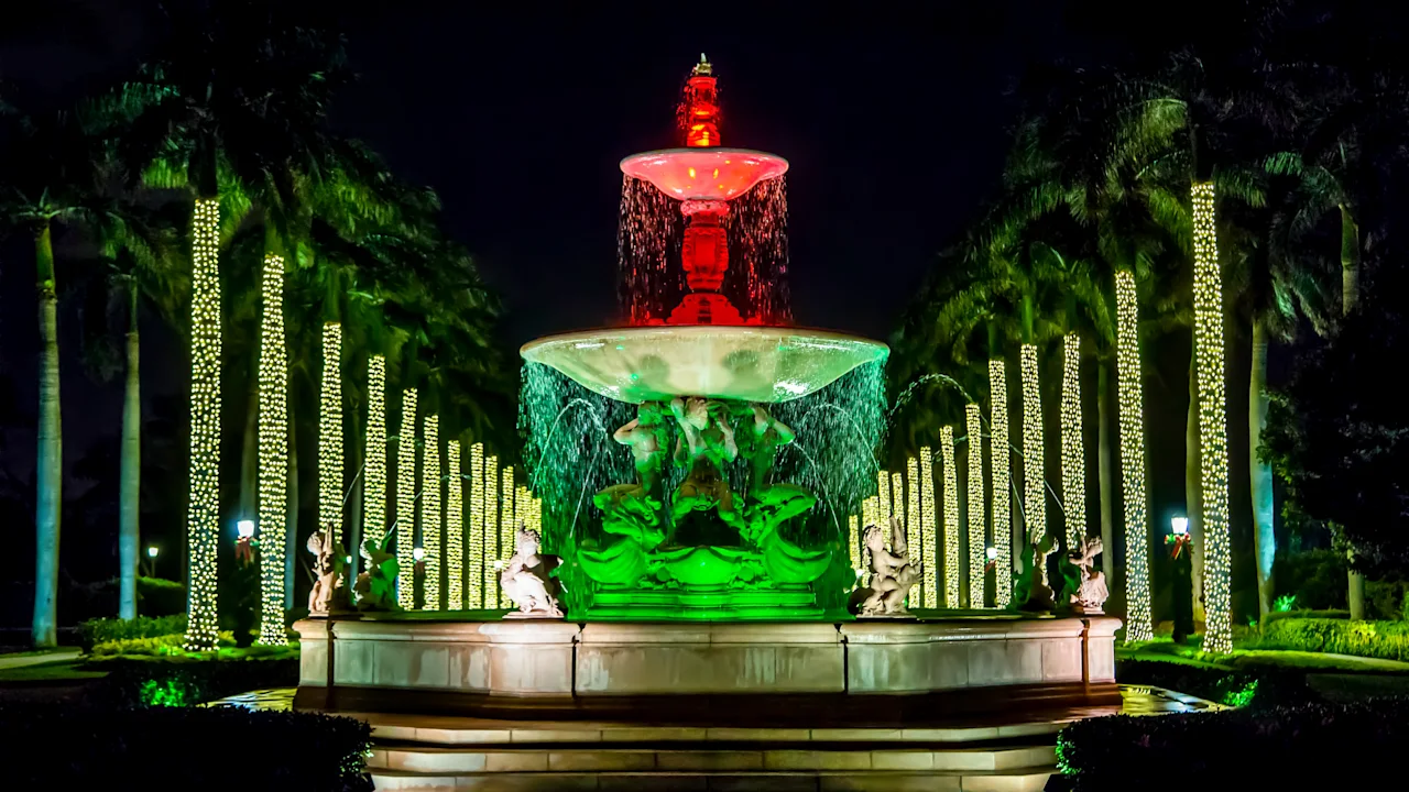 Illuminated fountain with red and green lights at night, surrounded by palm trees wrapped in lights.