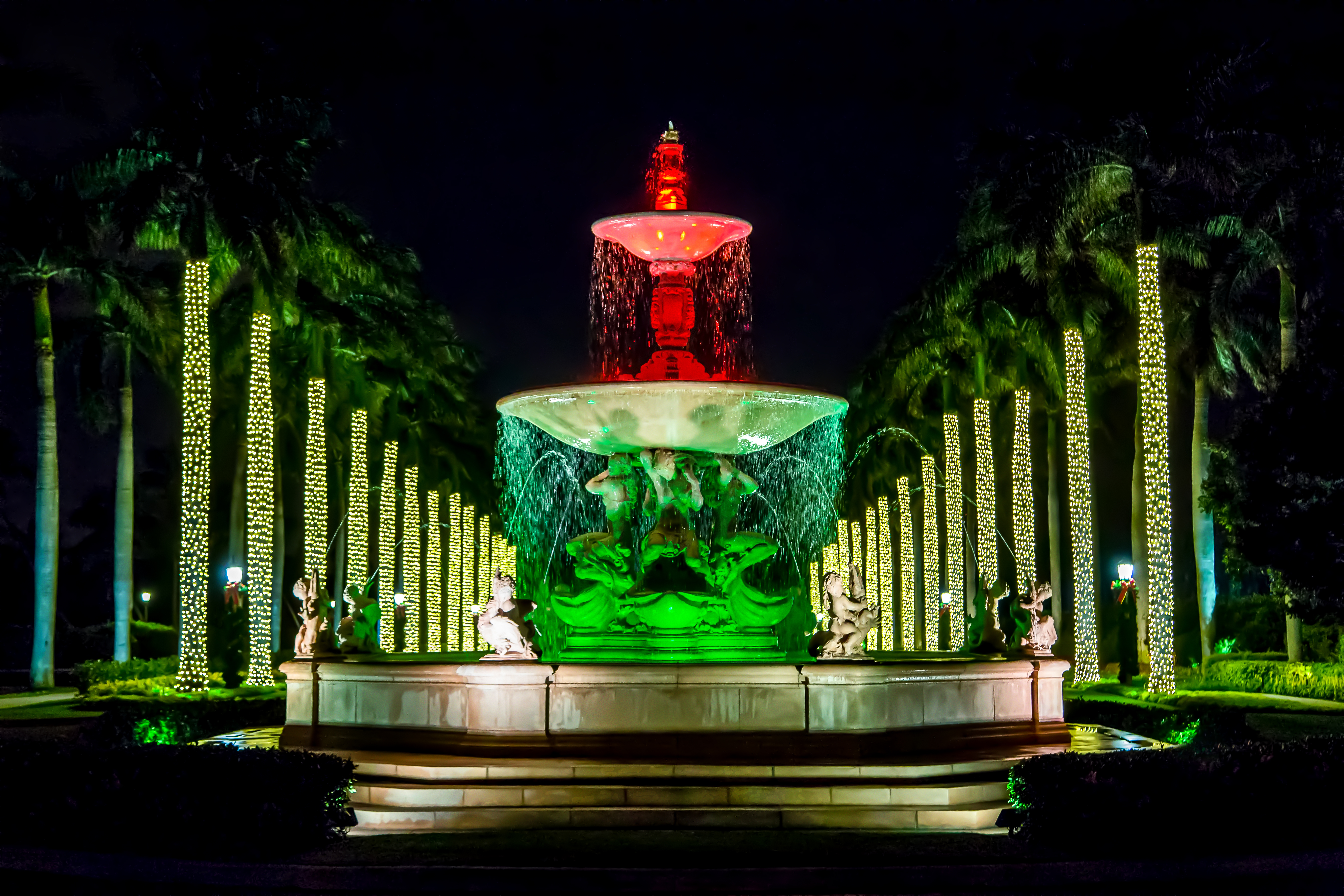Illuminated fountain at night with red and green lights, surrounded by palm trees wrapped in yellow lights.