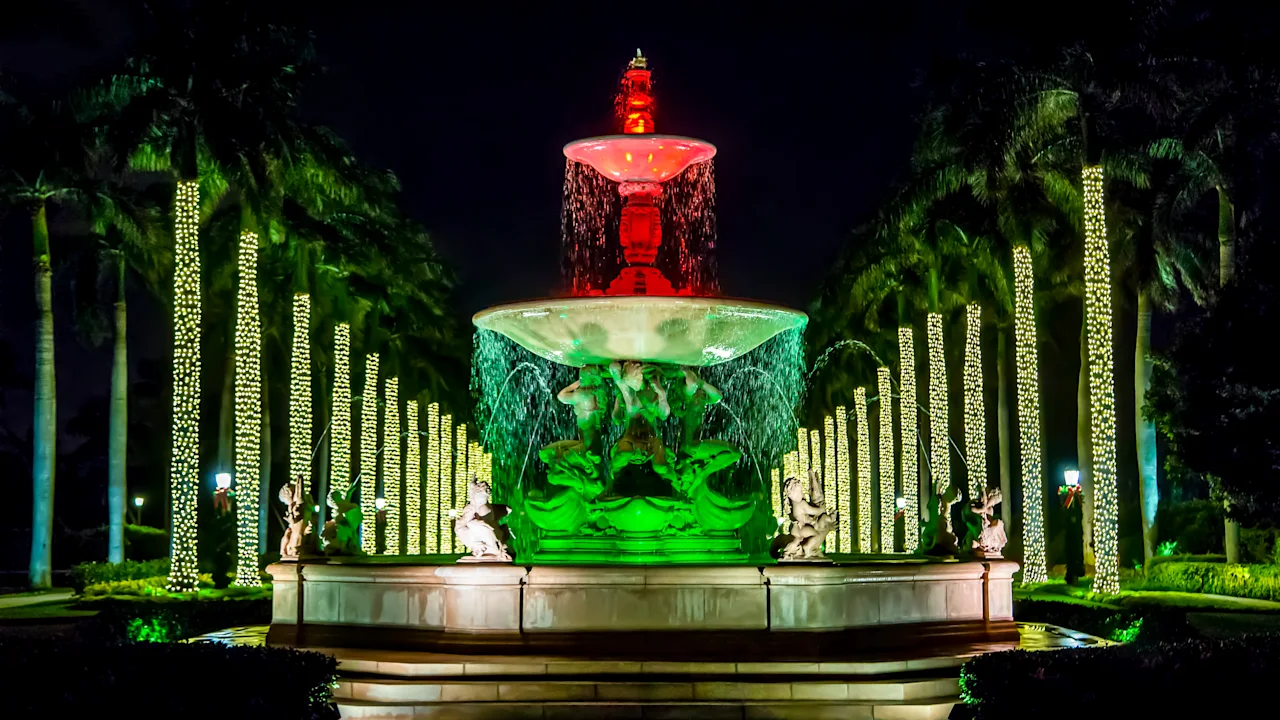 Illuminated fountain at night with red and green lights, surrounded by palm trees wrapped in yellow lights.