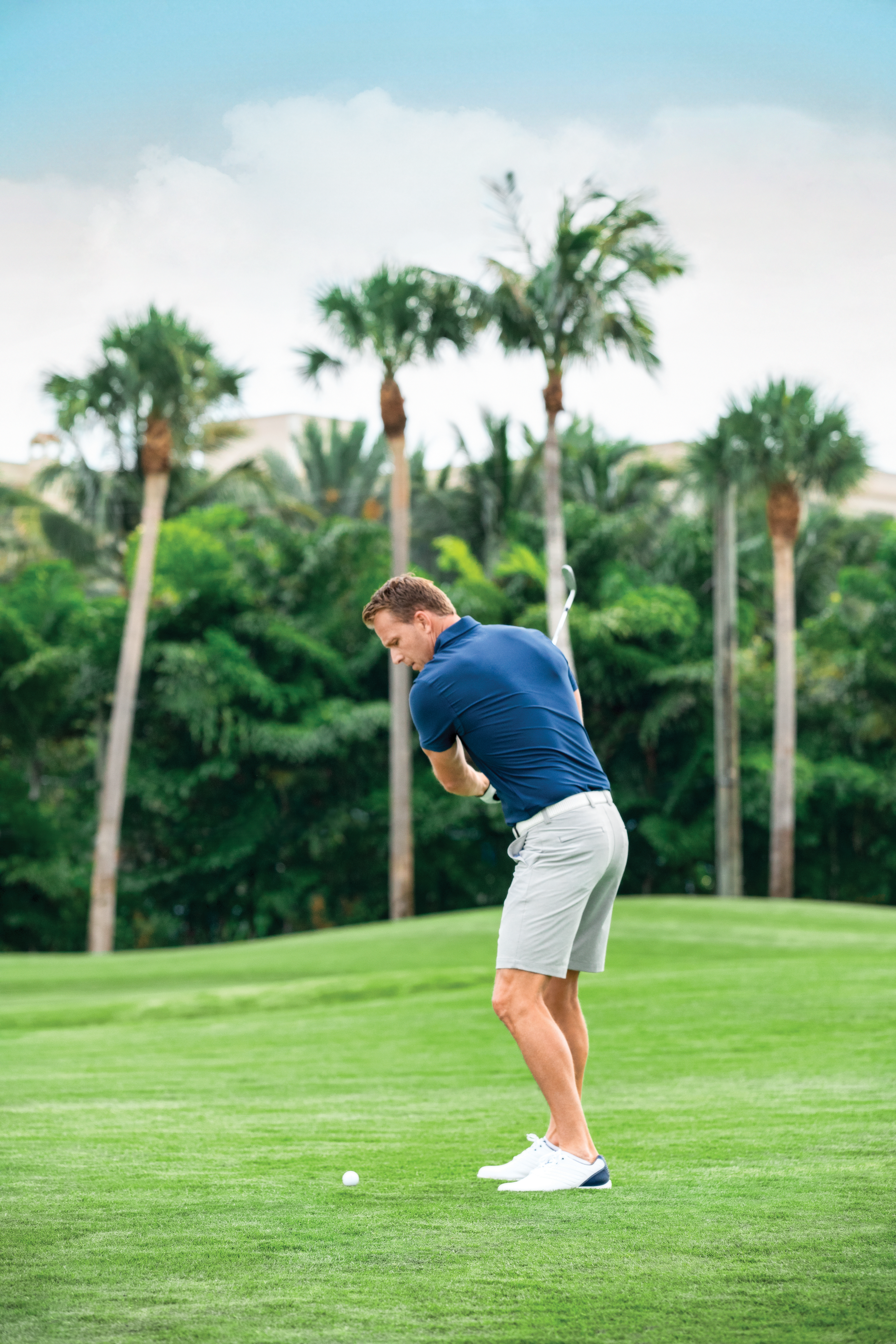 Man in blue shirt and white shorts swinging golf club on green course with palm trees in background.