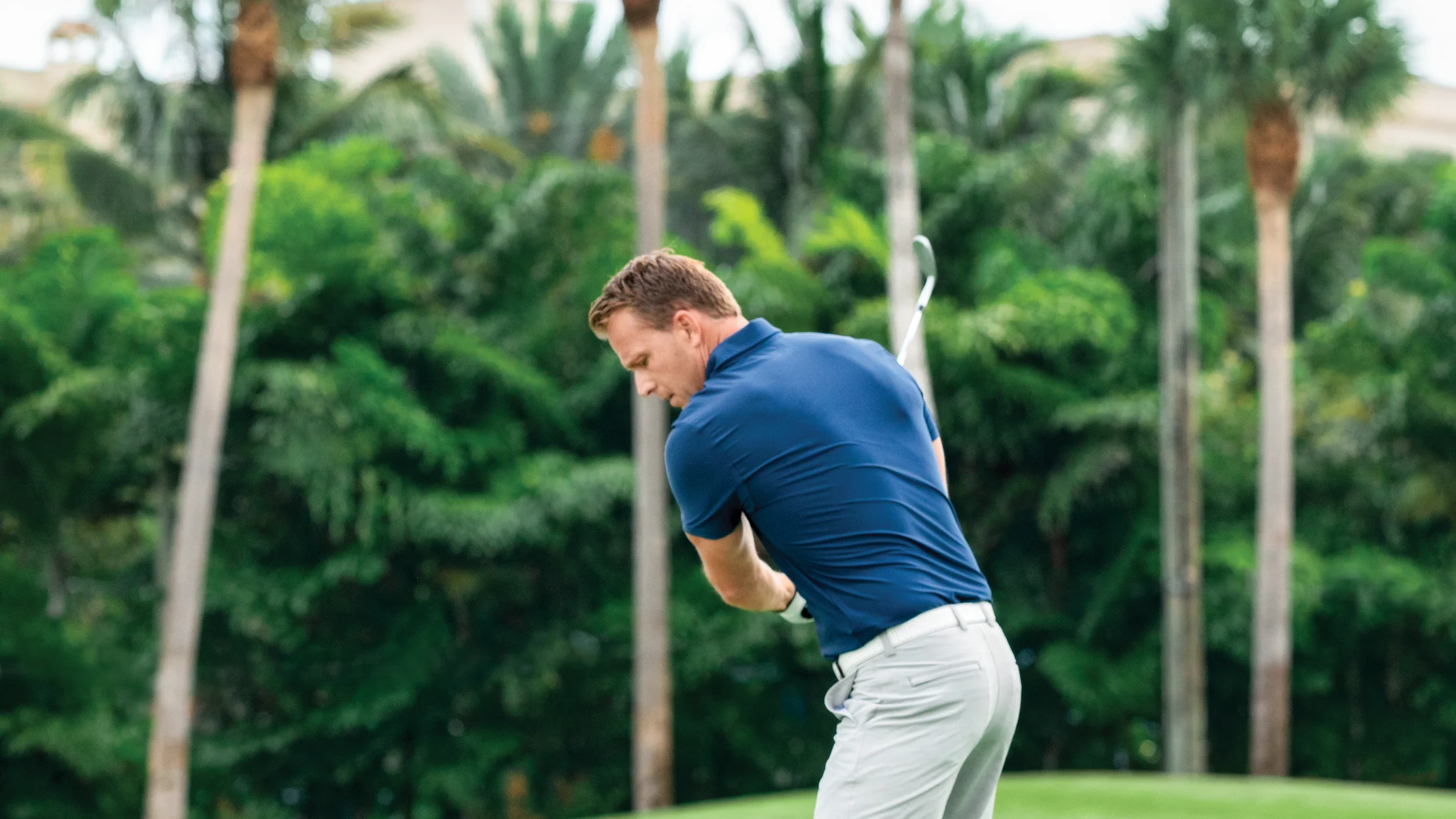 Man in blue shirt and white shorts swinging golf club on green course with palm trees in background.