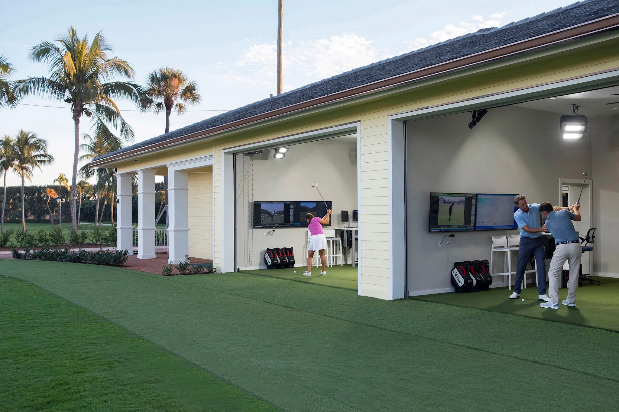 Golfers practicing at outdoor simulator bays with screens and palm trees in background.