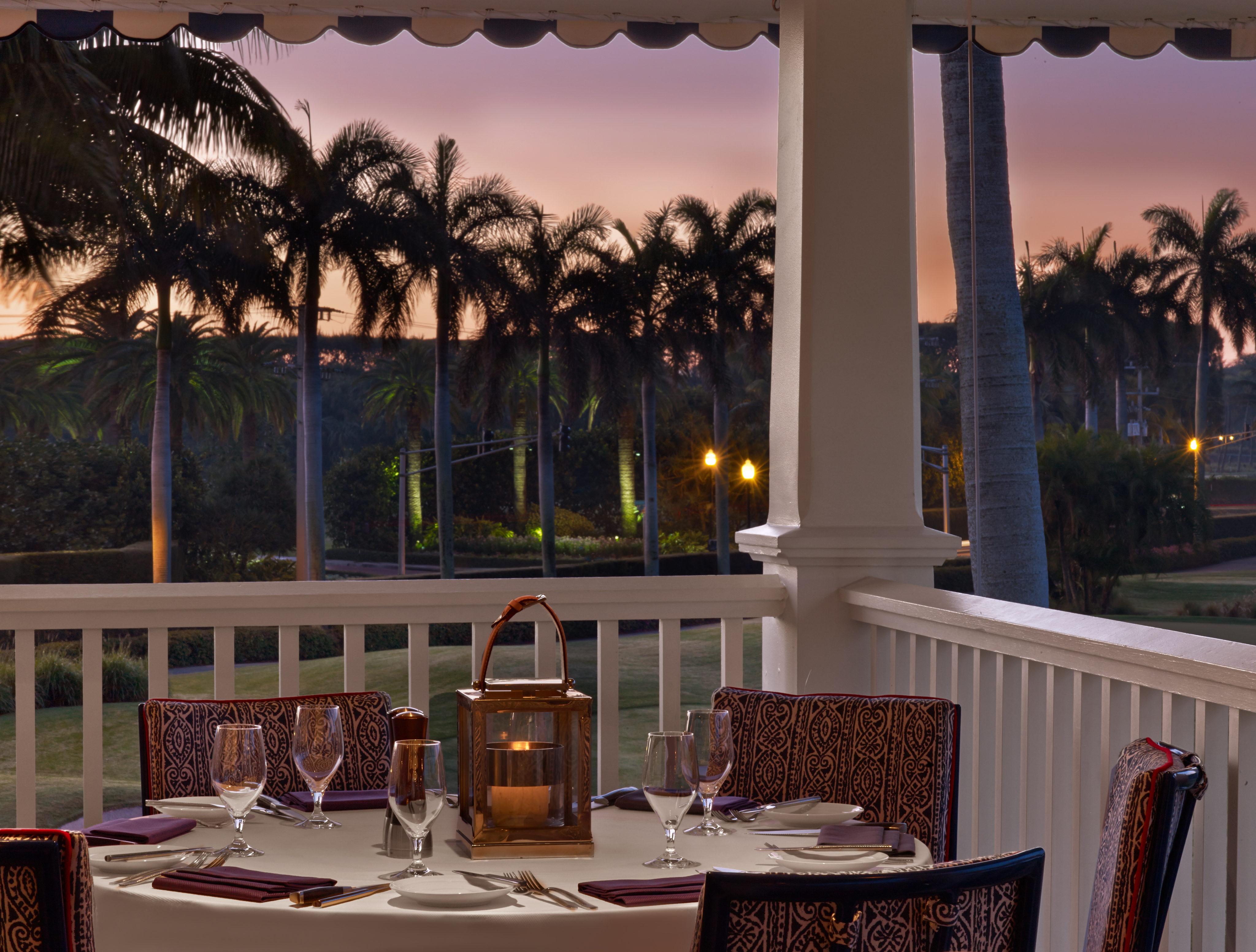 Elegant outdoor dining table set on a porch at sunset with palm trees and purple sky in background.