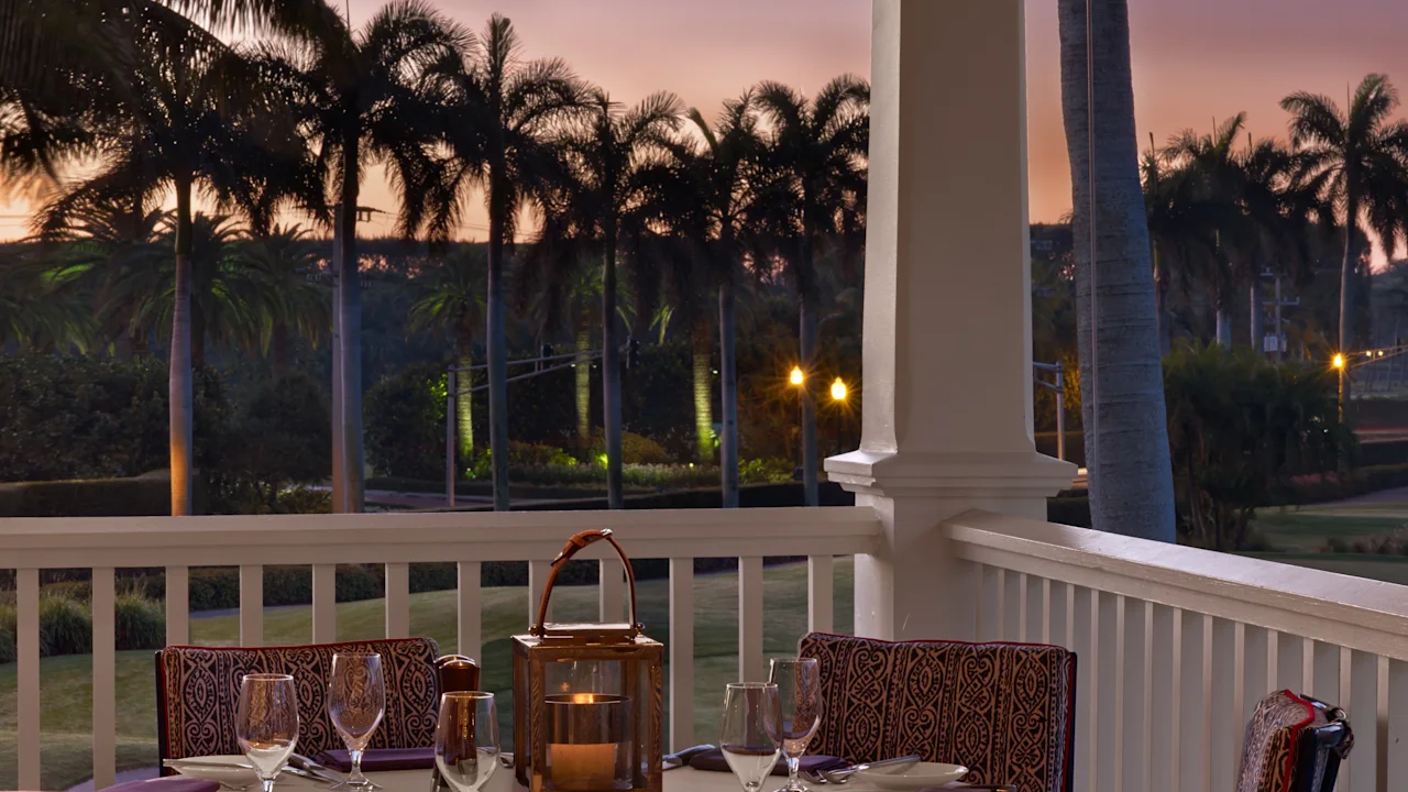 Elegant outdoor dining table set on a porch at sunset with palm trees and purple sky in background.
