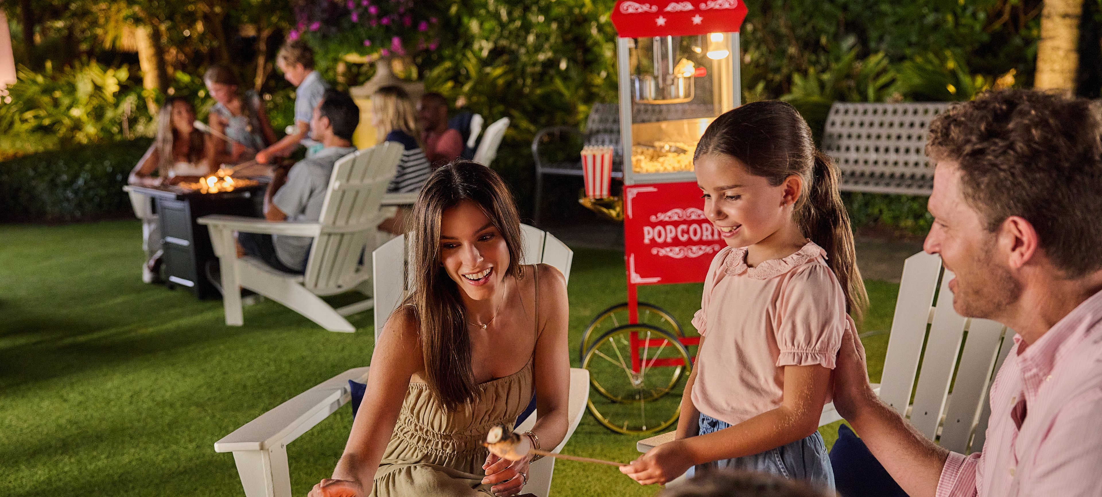 Family roasting marshmallows around outdoor fire pit with red popcorn machine in background.