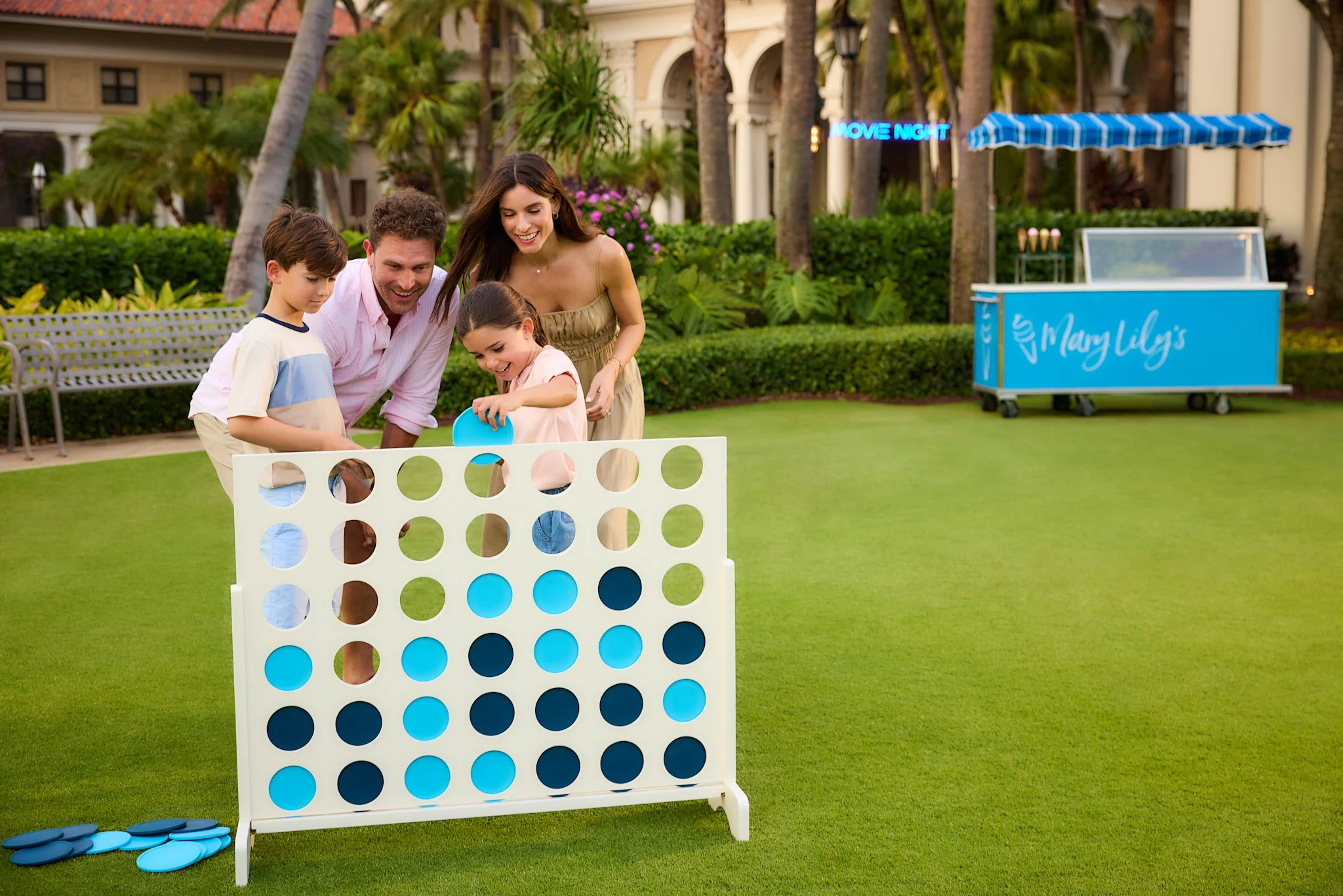 Family of four playing giant Connect Four game on resort lawn with blue ice cream cart in background.