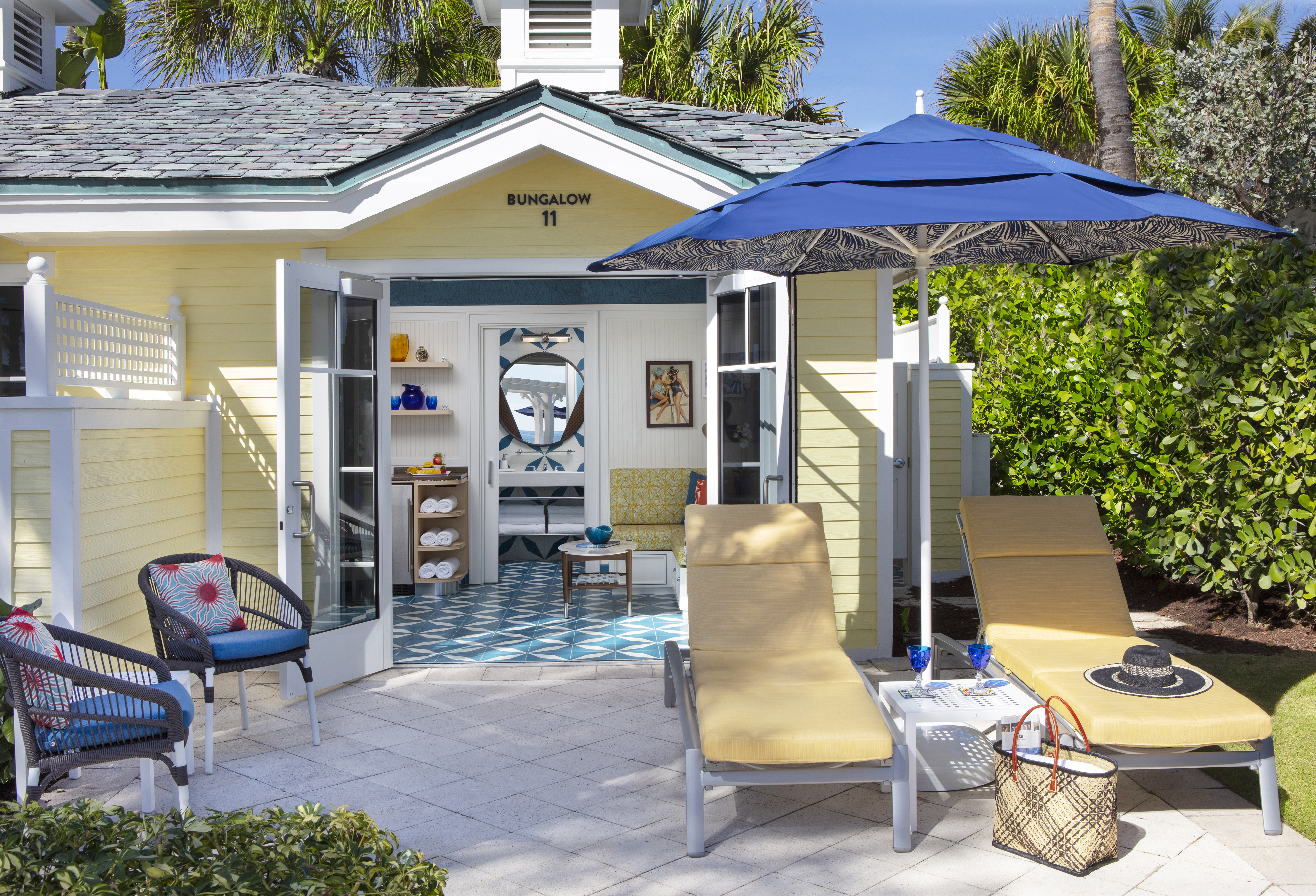 Yellow beach bungalow with blue umbrella, lounge chairs, and patterned tile floor visible inside.