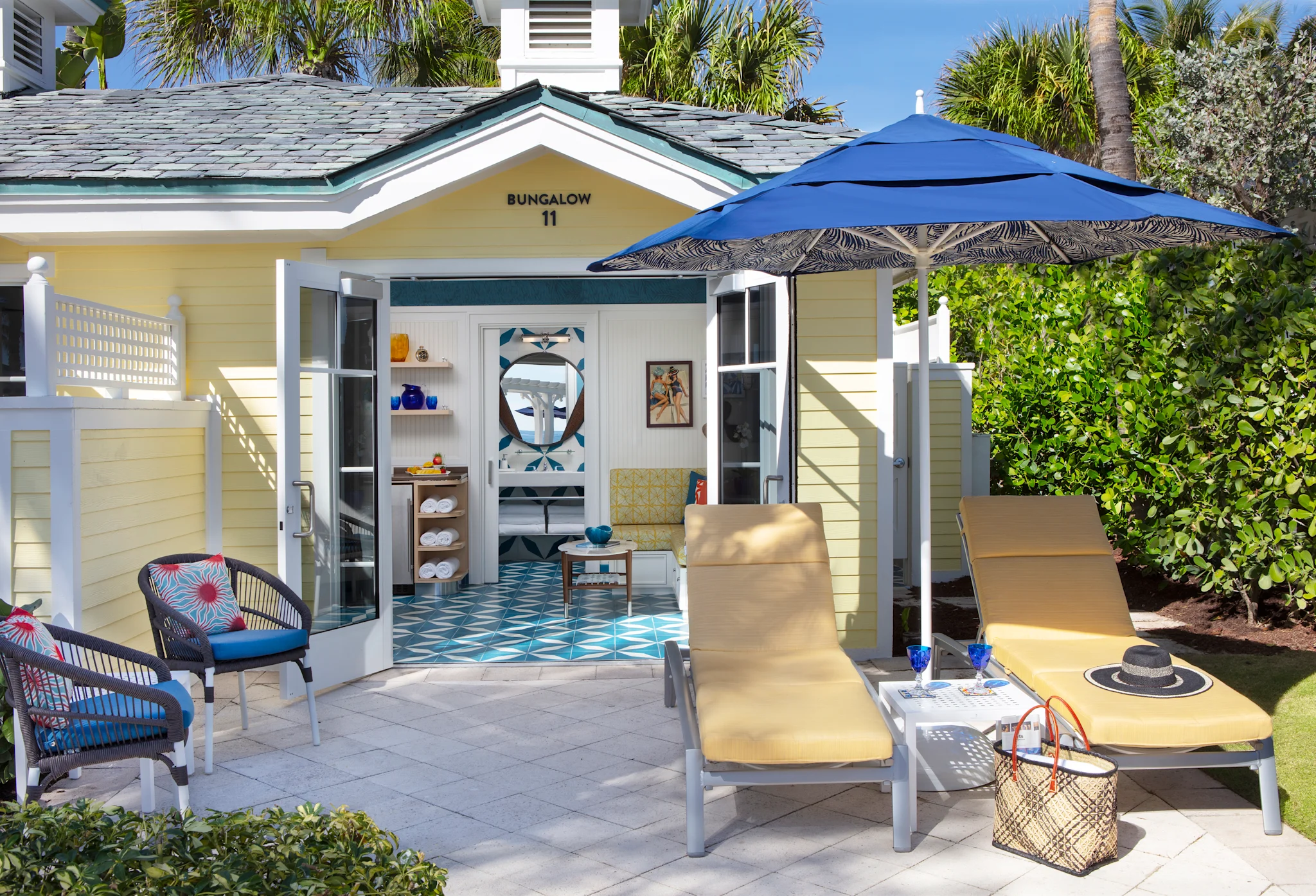 Yellow beach bungalow with blue umbrella, lounge chairs, and patterned tile floor visible inside.
