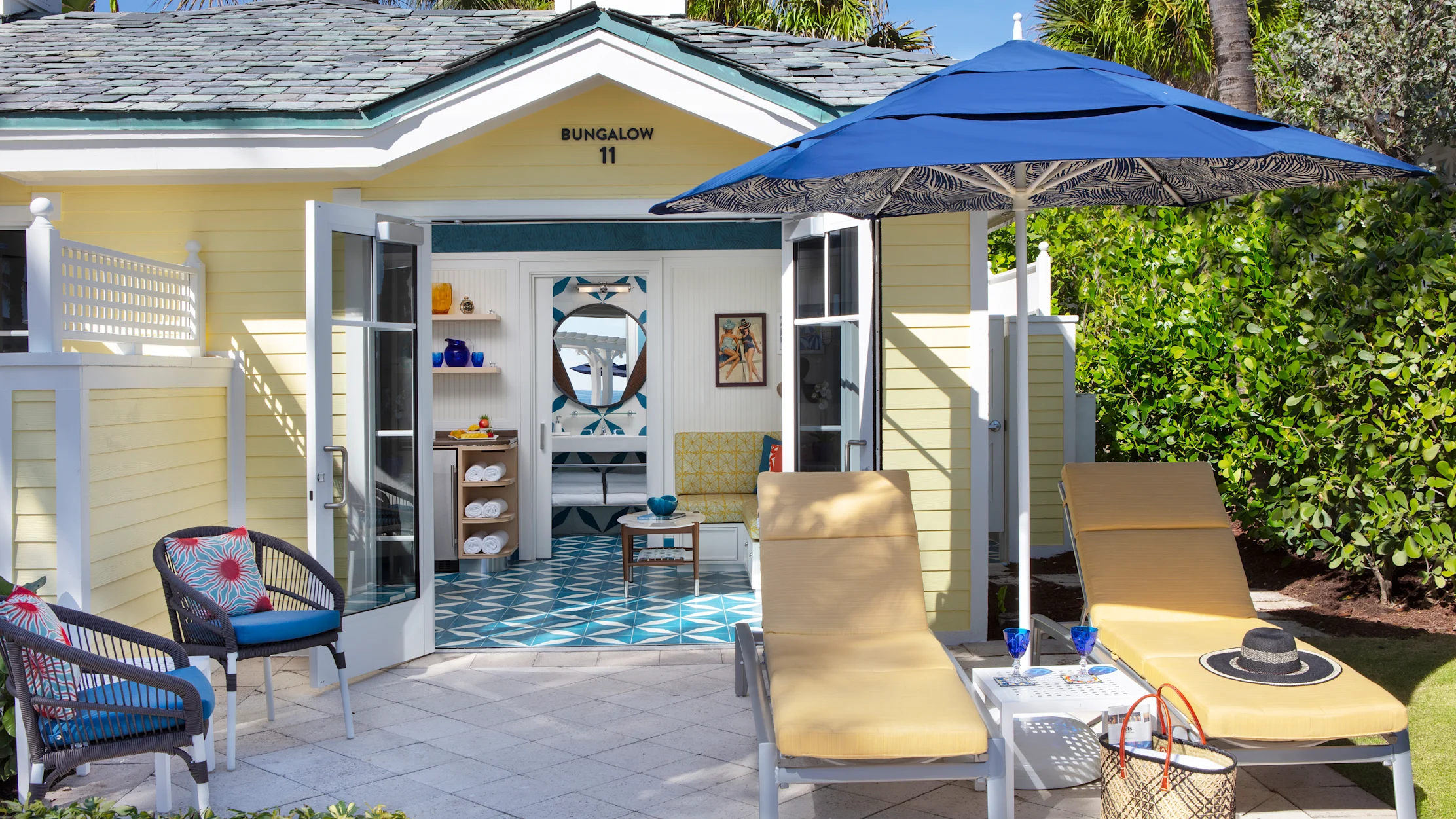 Yellow beach bungalow with blue umbrella, lounge chairs, and patterned tile floor visible inside.