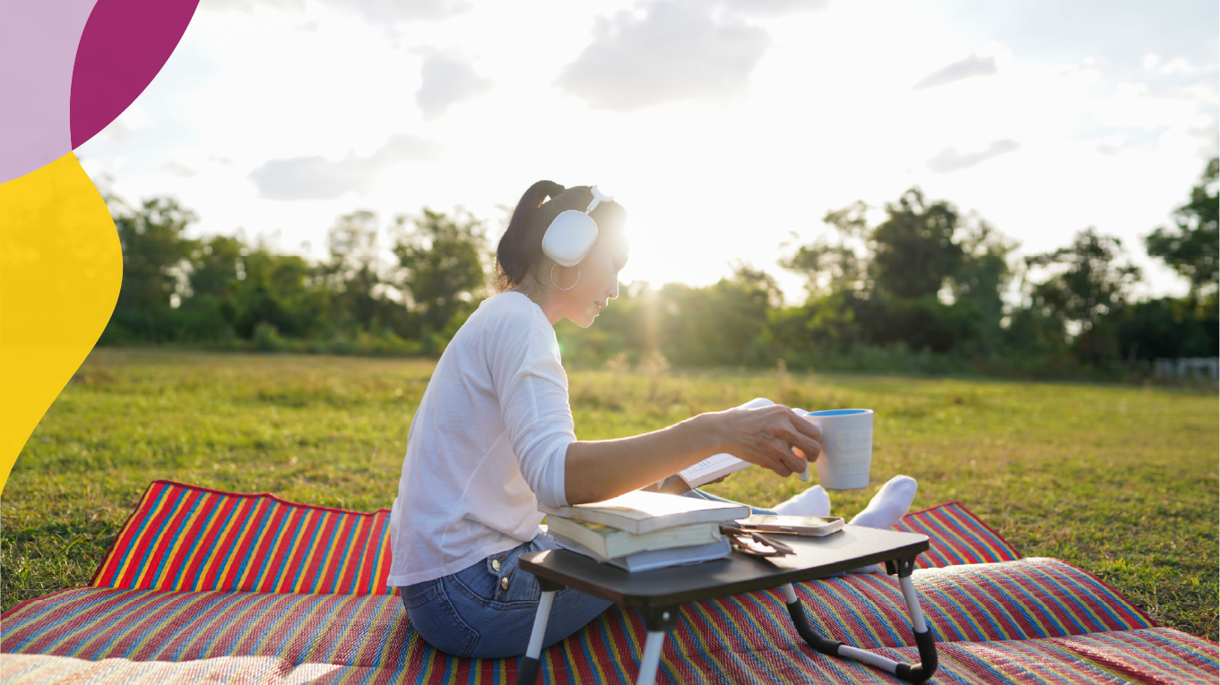 Woman sitting on a blanket in a park reading with headphones on and a cup of coffee.