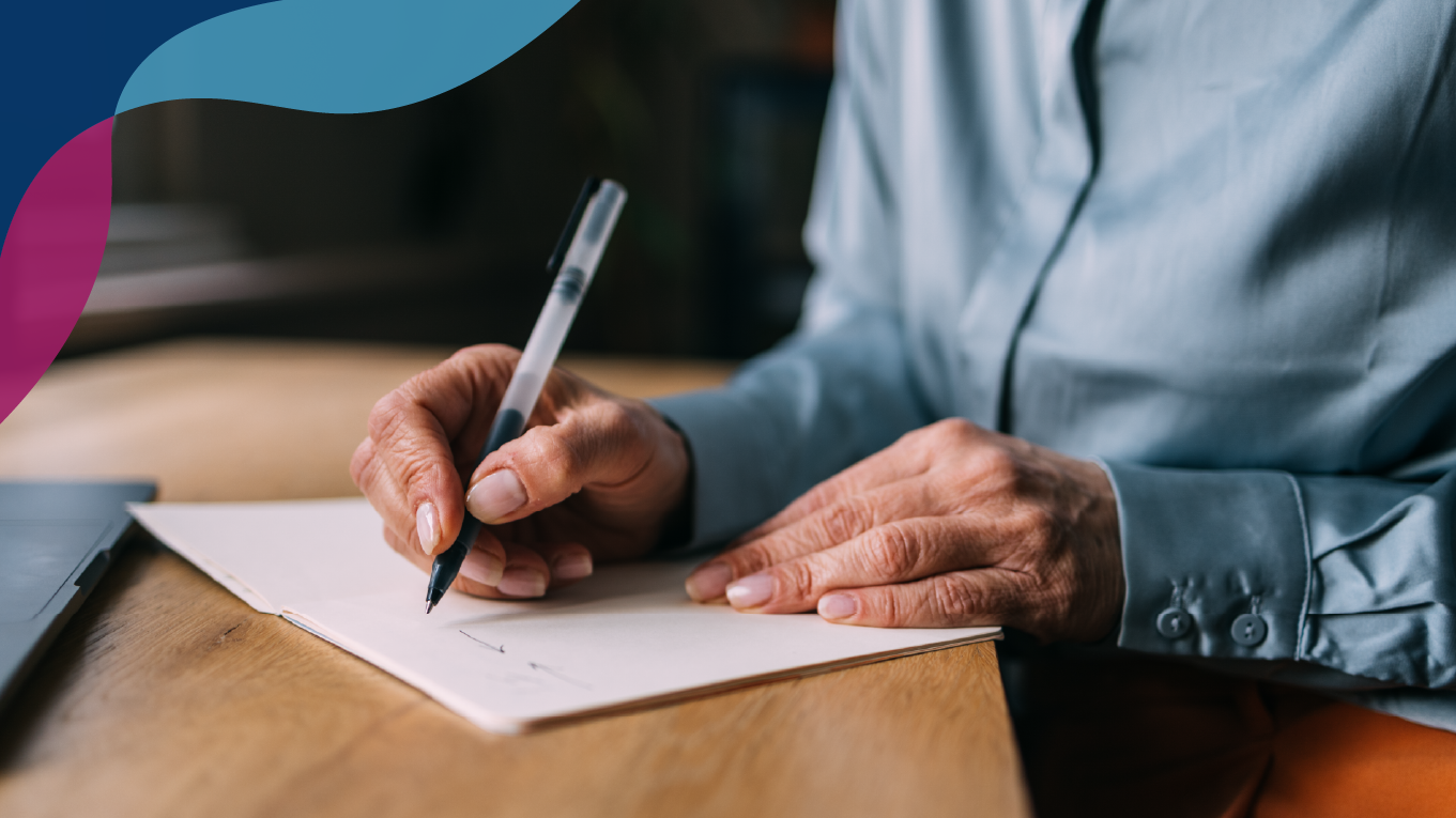 Person in a light blue silk long sleeve top, writing in a notebook on top of a wooden table.