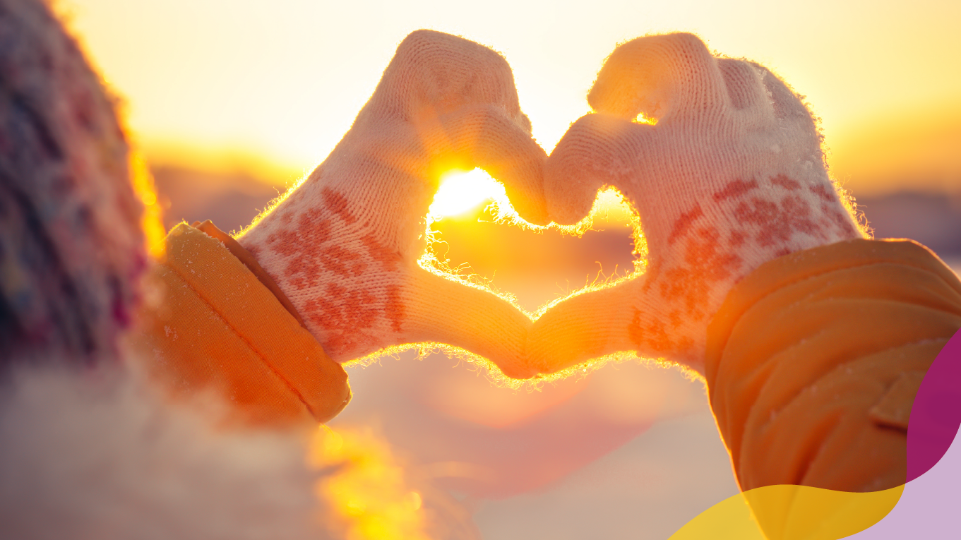 A person standing outside, surrounded by a snowy landscape, wearing gloves and making a heart shape with hands.