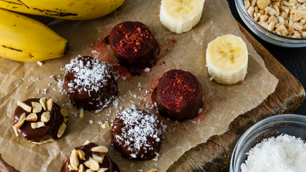 Chocolate-covered bananas with nuts and coconut toppings on a cutting board.