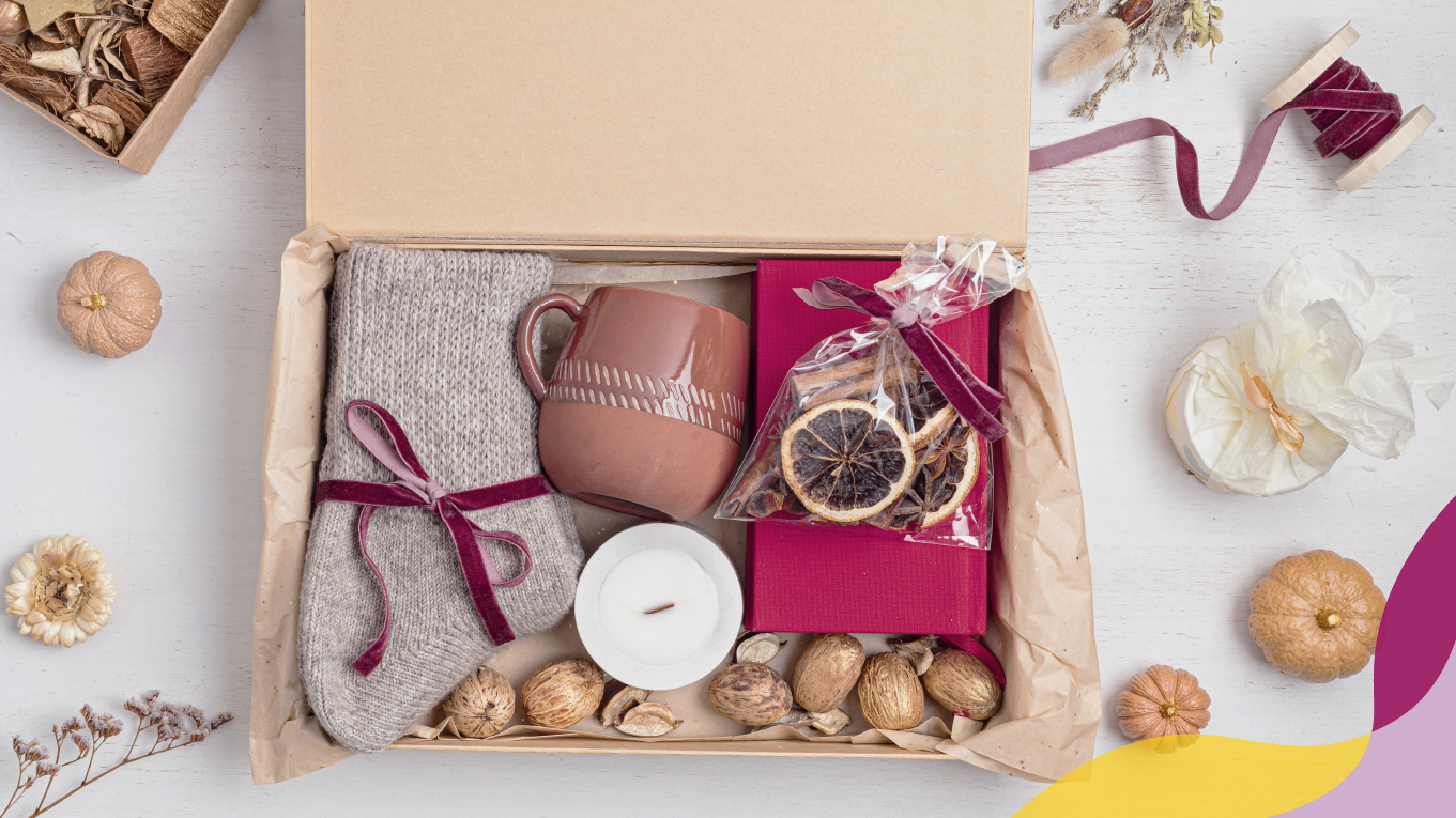 Gift box filled with a pair of gray/brown socks, a brown/tan mug, a white candle, and a magenta journal.