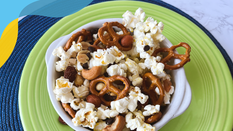 Green plate with a white bowl filled with nuts, popcorn, and pretzels.