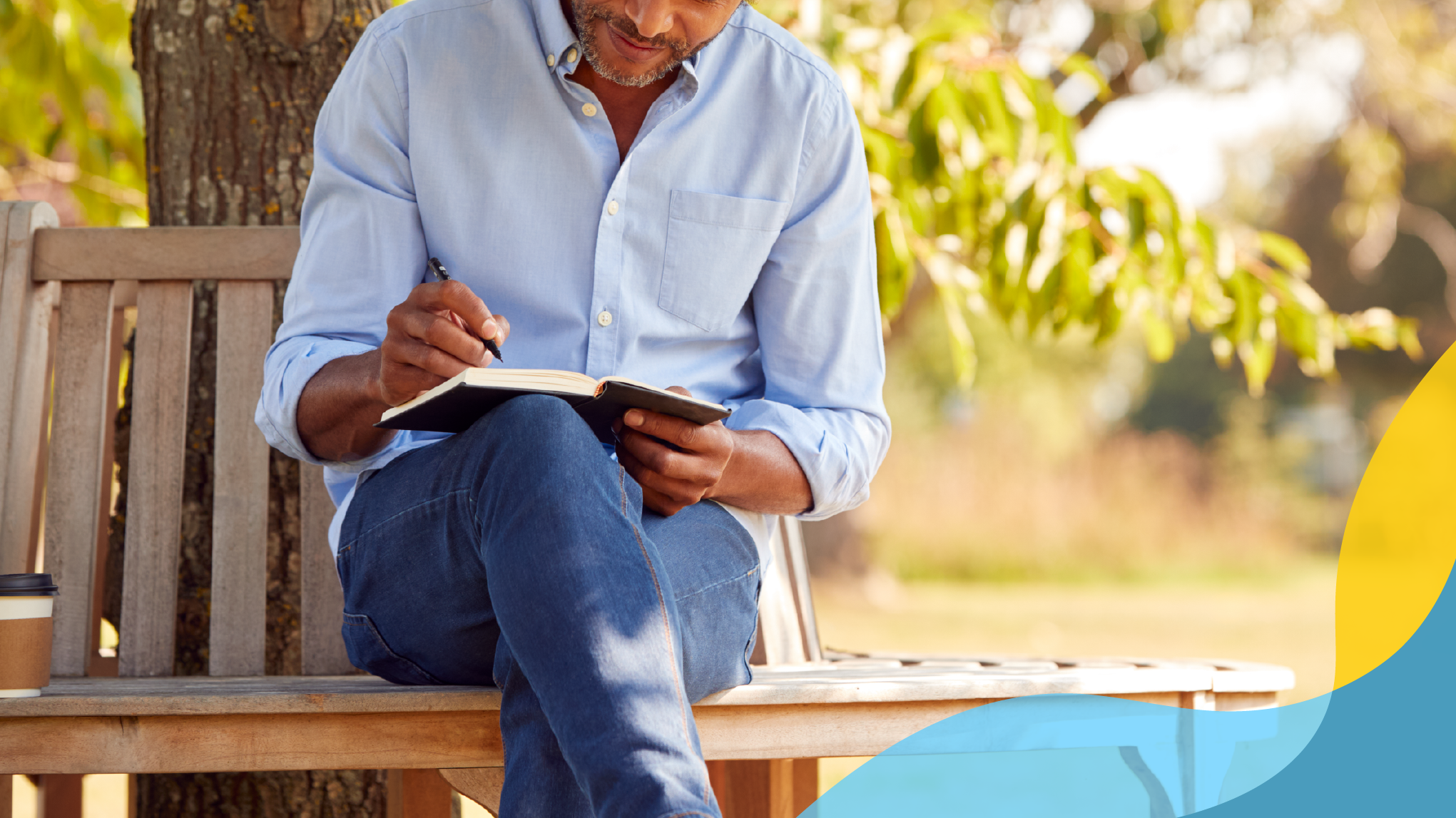 Black man sitting on a park bench writing in a journal on a sunny day.