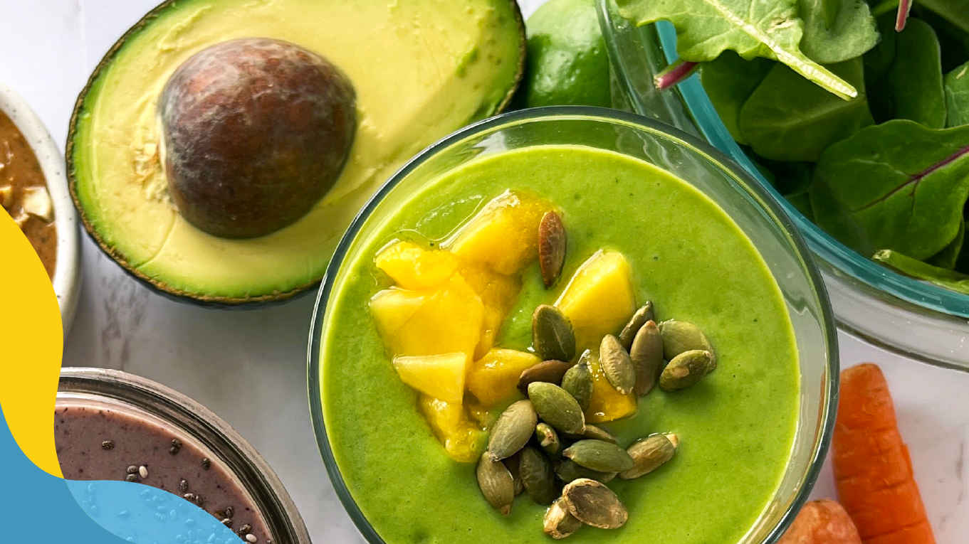 Top view of a green smoothie surrounded by an avocado cut in half, a bowl of fresh greens, and carrots.