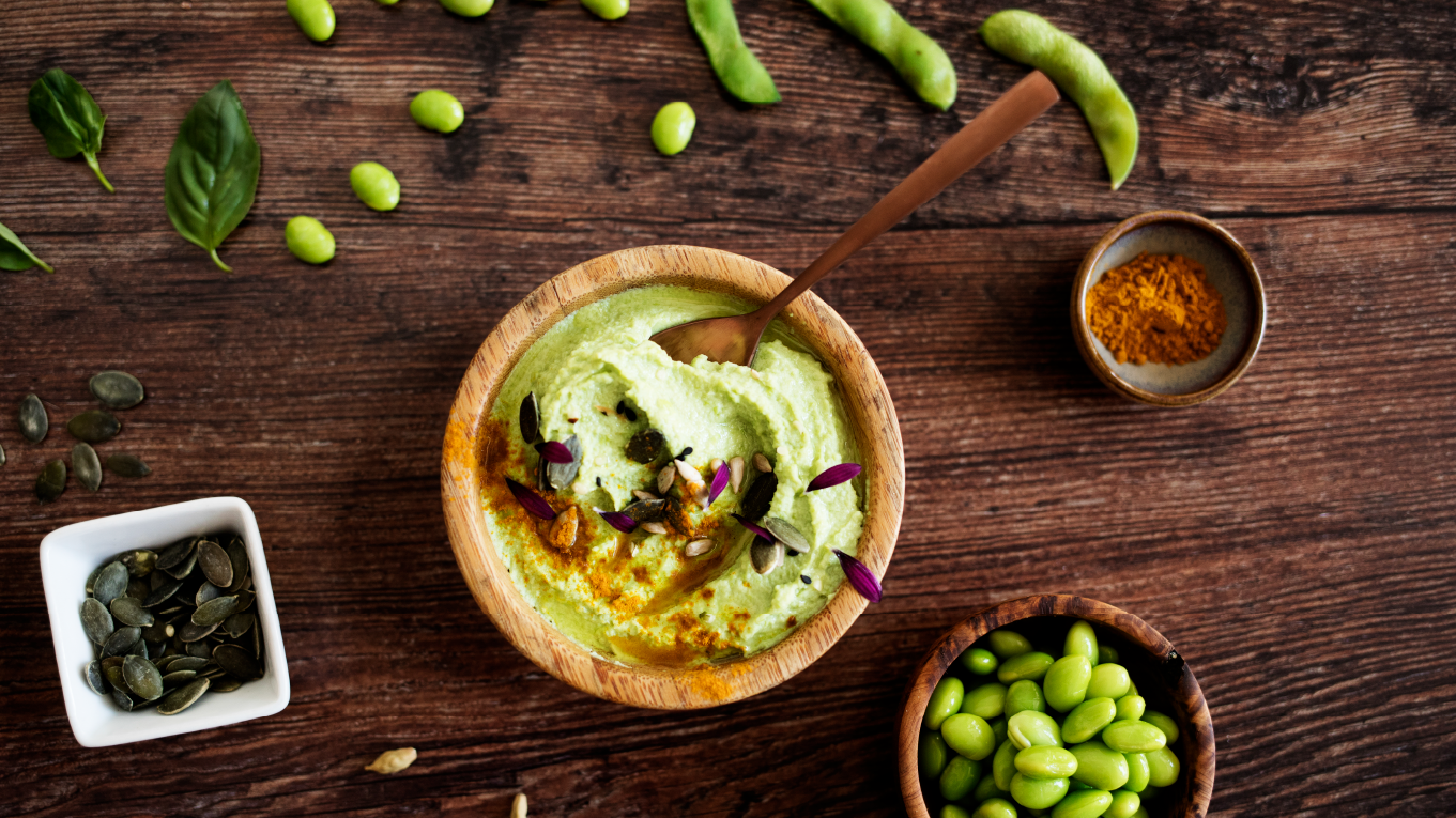 A wooden bowl of edamame guacamole, next to the main wooden bowl is a smaller bowl filled with edamame and several spices surrounding it.