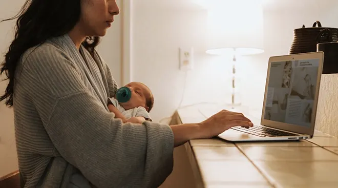 mother working on computer at desk while holding baby in arms