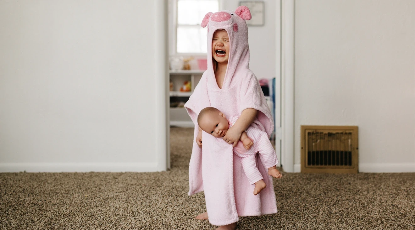 crying three year old wearing pink towel and holding baby doll at home
