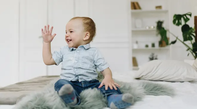 toddler sitting on bed waving hello