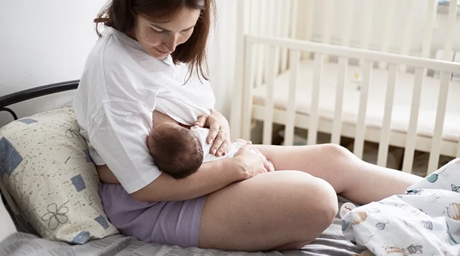 mother breastfeeding baby while sitting on bed at home