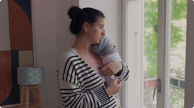 A postpartum mother holding her newborn baby while looking out the window at home. 