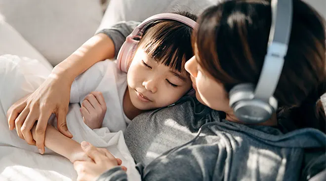 Young girl and her mom listen to podcast on headphones while laying in bed.