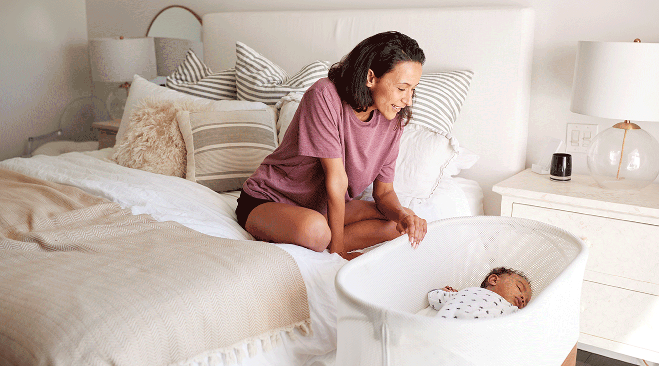 mother looking at baby sleeping in bassinet next to bed at home