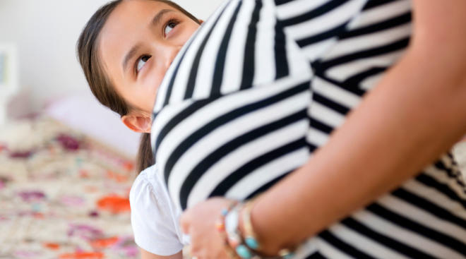 Pregnant woman standing with her older child peering at her pregnant stomach.