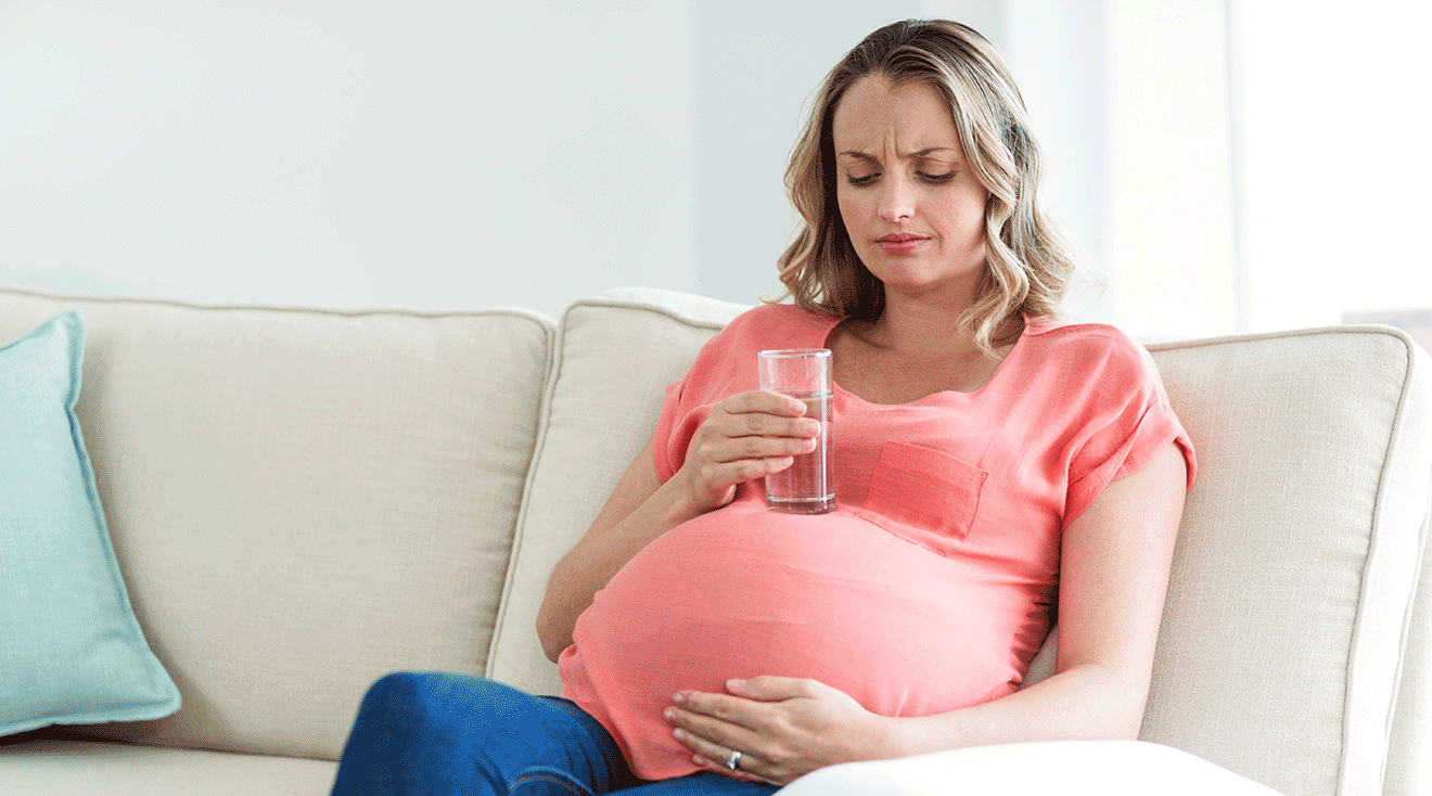 cpnpregnant woman drinking a glass of water while sitting on the couch at home