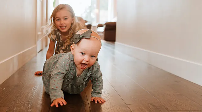 Big sister crawling behind baby sister down hallway at home.