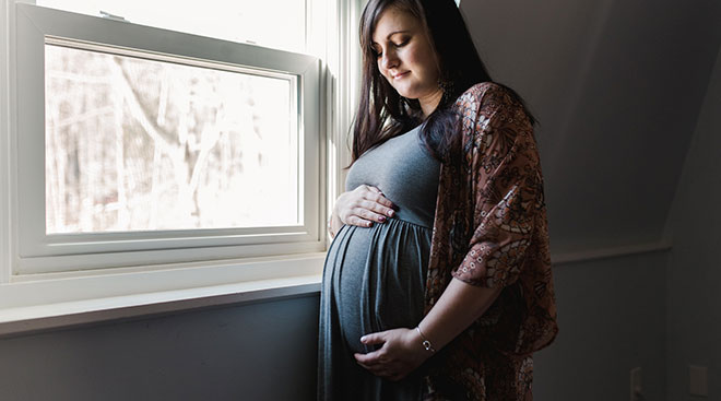 serious pregnant woman touching her belly at home by the window