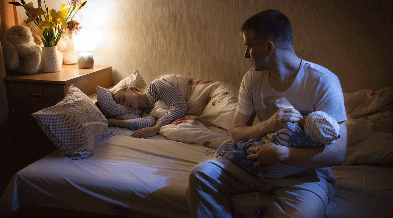 dad feeding baby at night while mom sleeps in bed