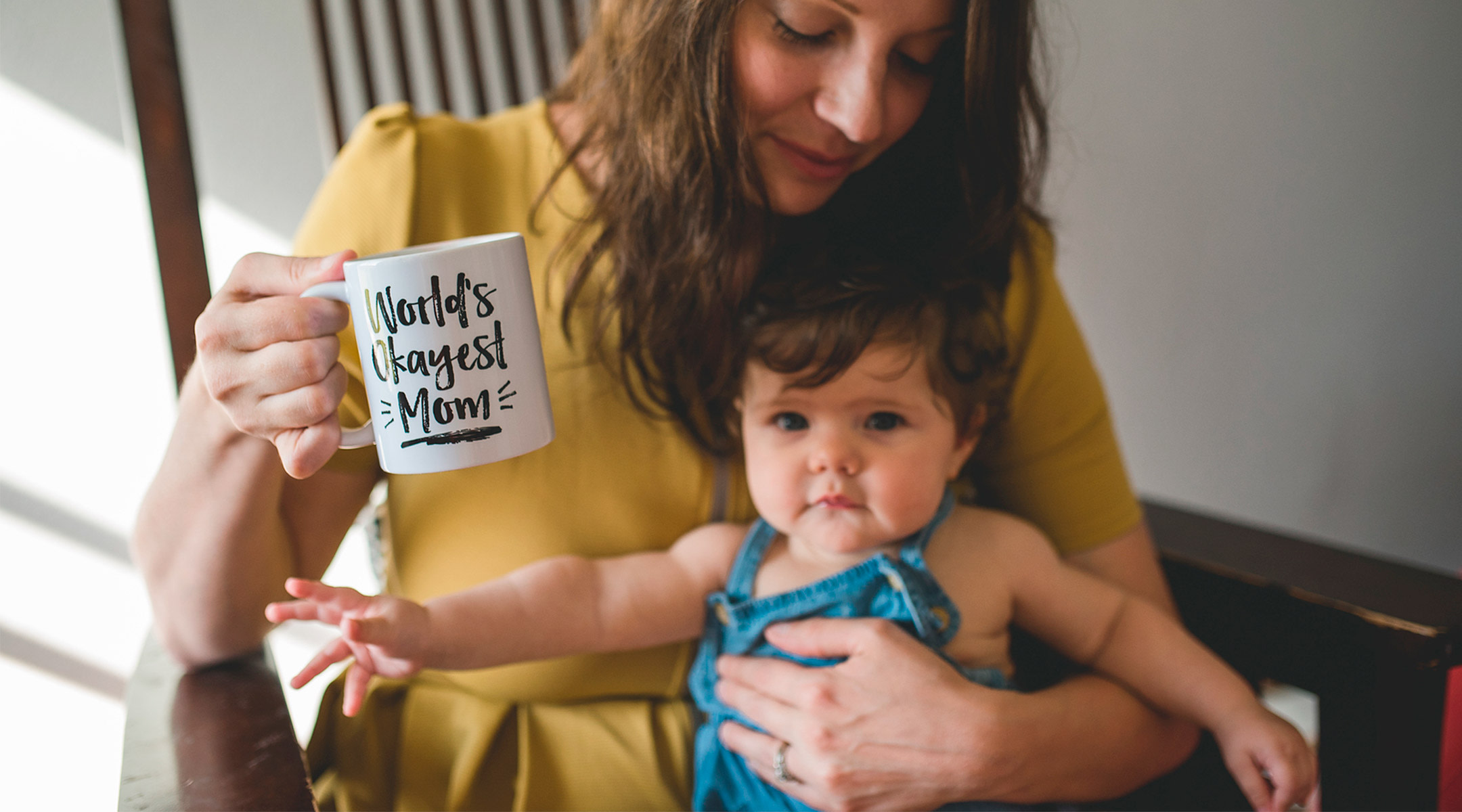 mom holding baby with mug that says, "world's okayest mom" speaking to the idea of mom shaming