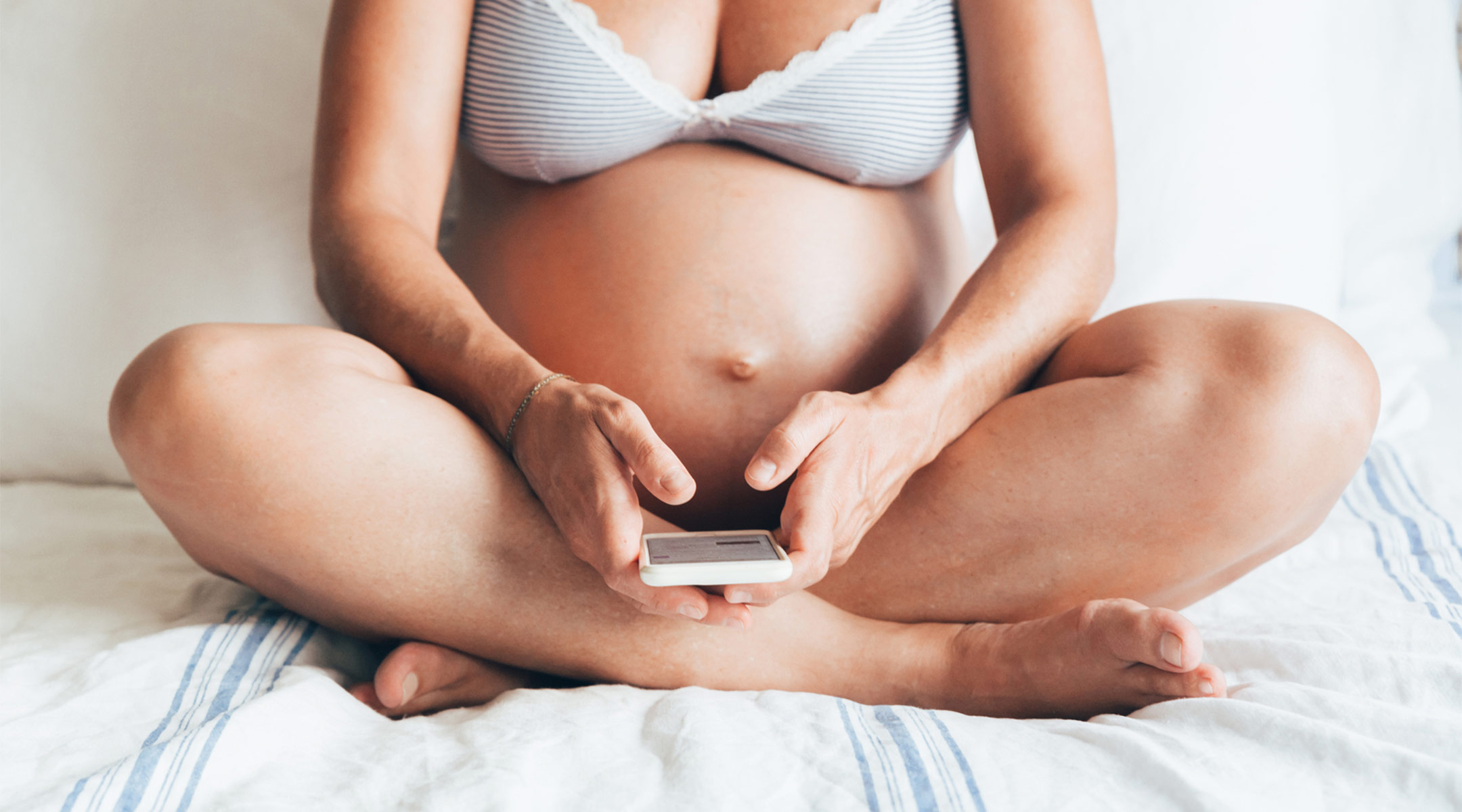 pregnant woman sitting on bed in her underwear with her phone