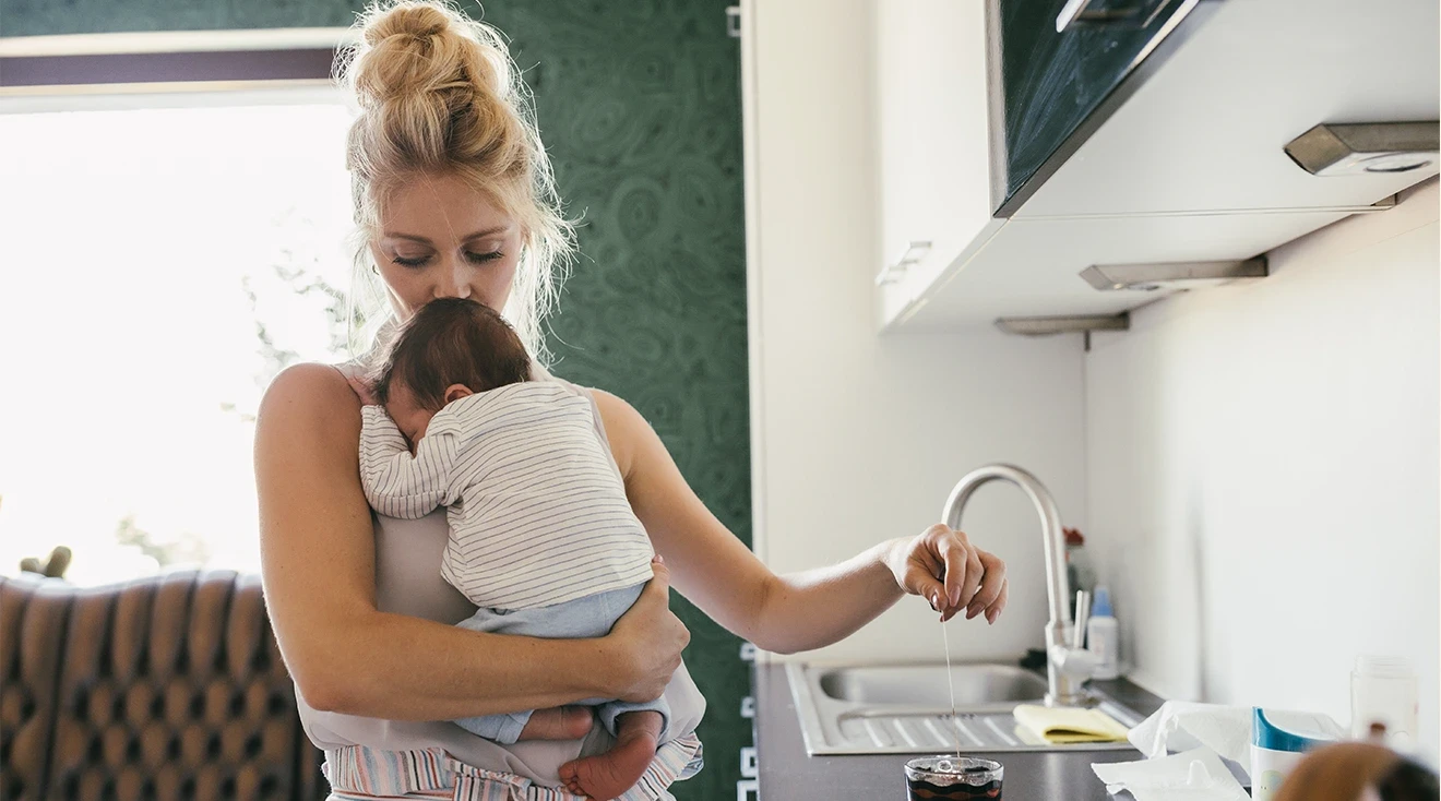 mother holding newborn baby while in the kitchen making tea