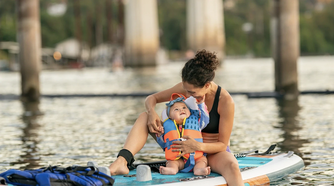 baby wearing a life jacket while sitting on paddle board in water with mom