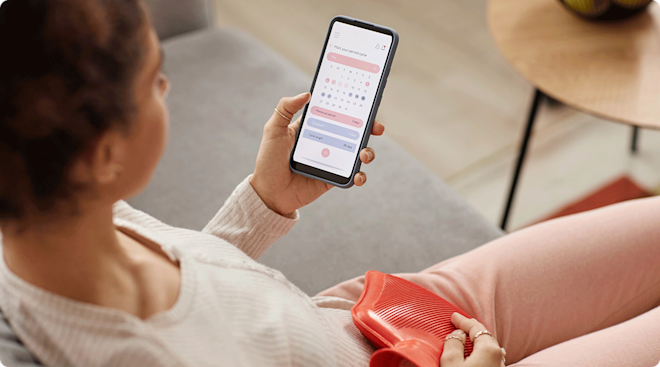 woman looking at period tracker calendar on phone while using a heating pad