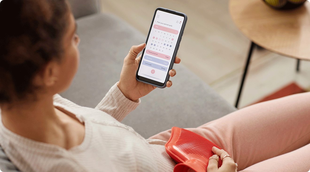 woman looking at period tracker calendar on phone while using a heating pad