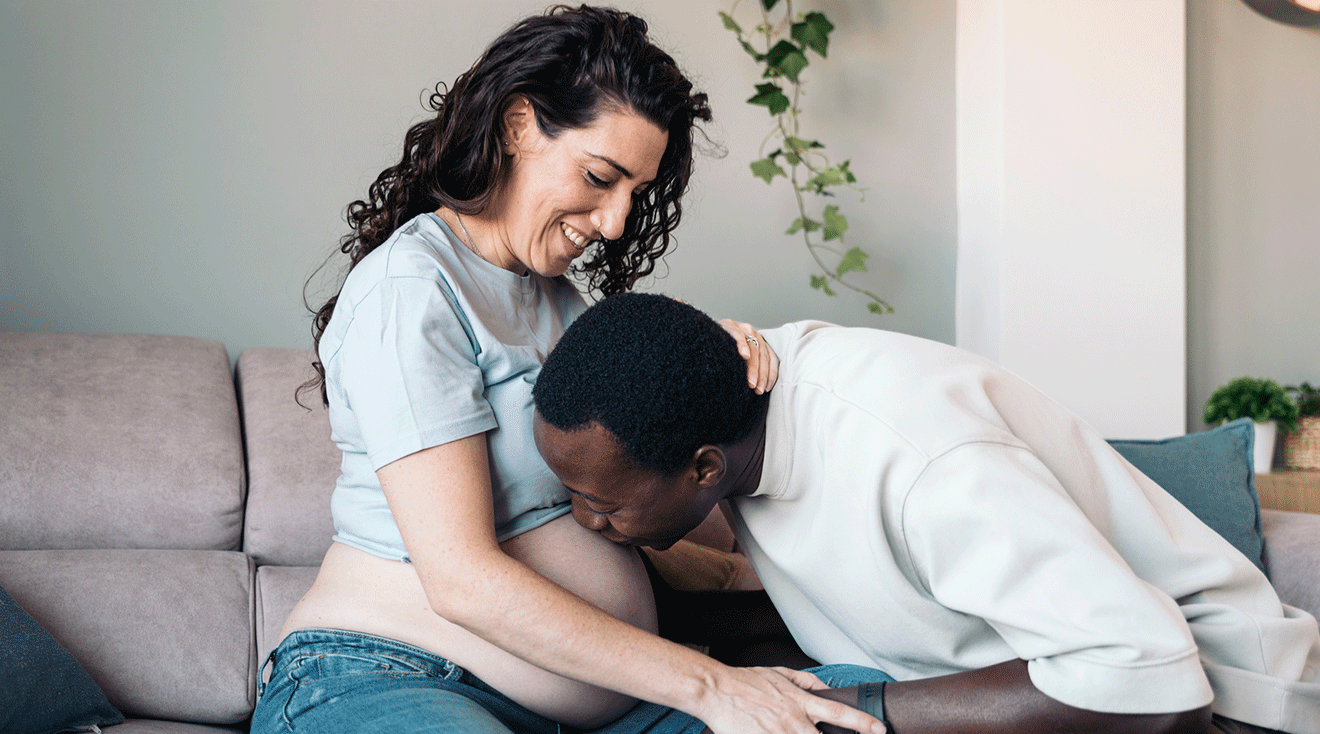 husband kissing wife's pregnant belly while sitting on couch at home