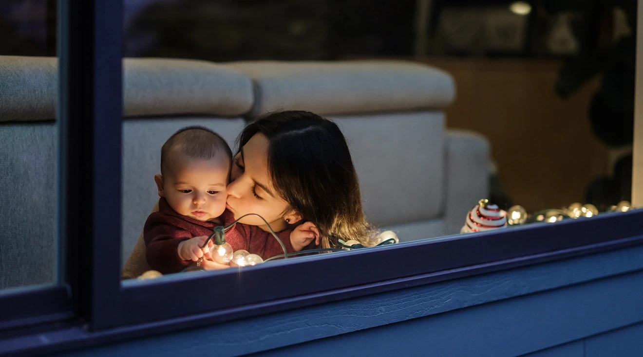 mother kissing baby white looking at christmas lights near the window of home